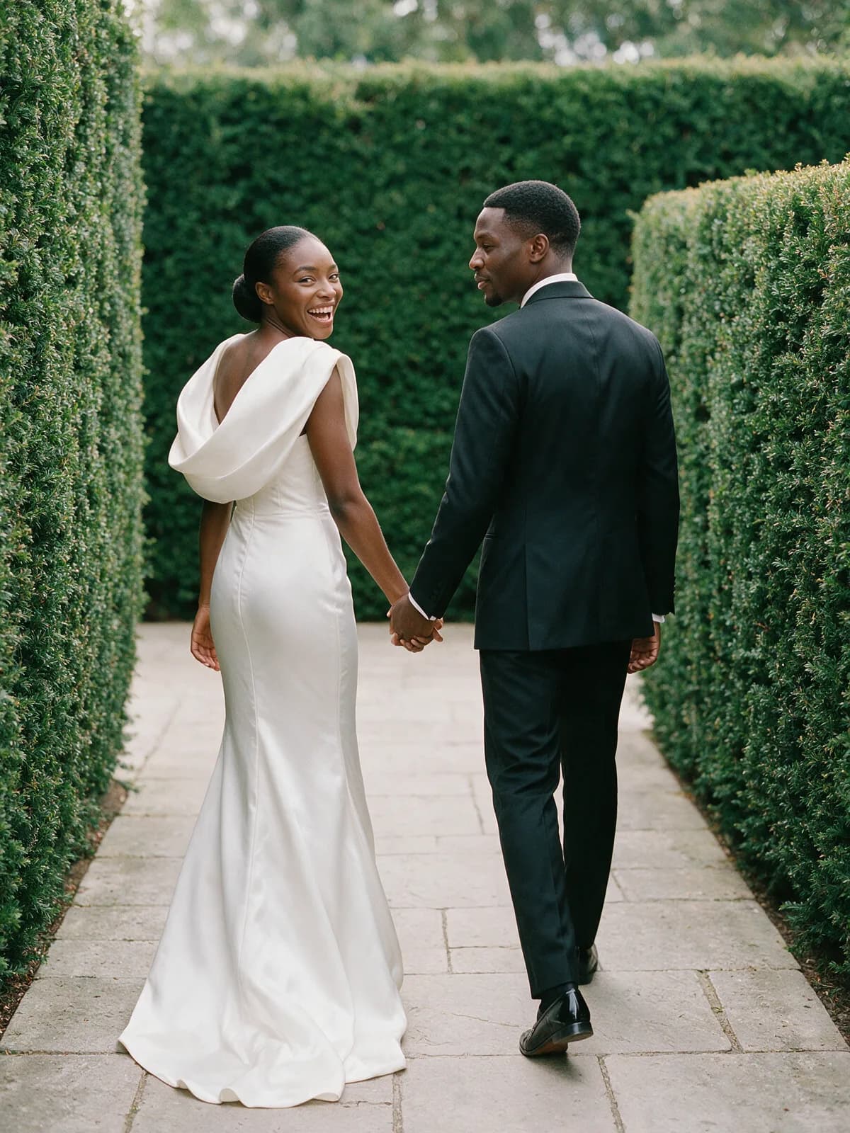 Mutinta glances back laughing as she and Kabwe walk hand in hand between tall hedges